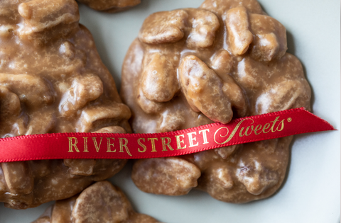 Two large golden pralines atop a white marble surface with a red ribbon draped across bearing the river street sweets logo in gold