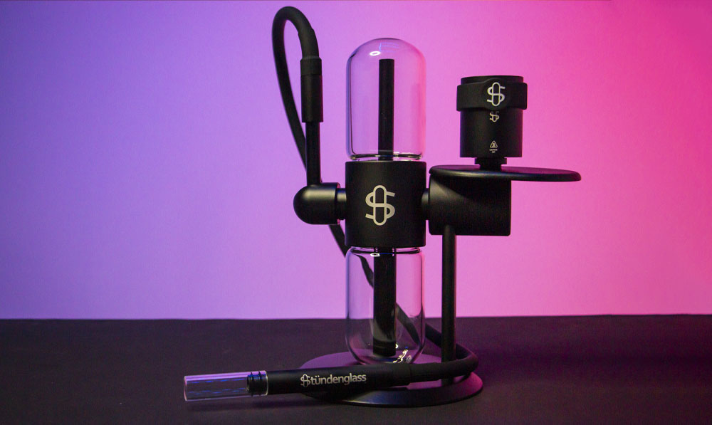 يقف Stundenglass Kompact على سطح أسود أمام خلفية استوديو باللونين الأرجواني والوردي