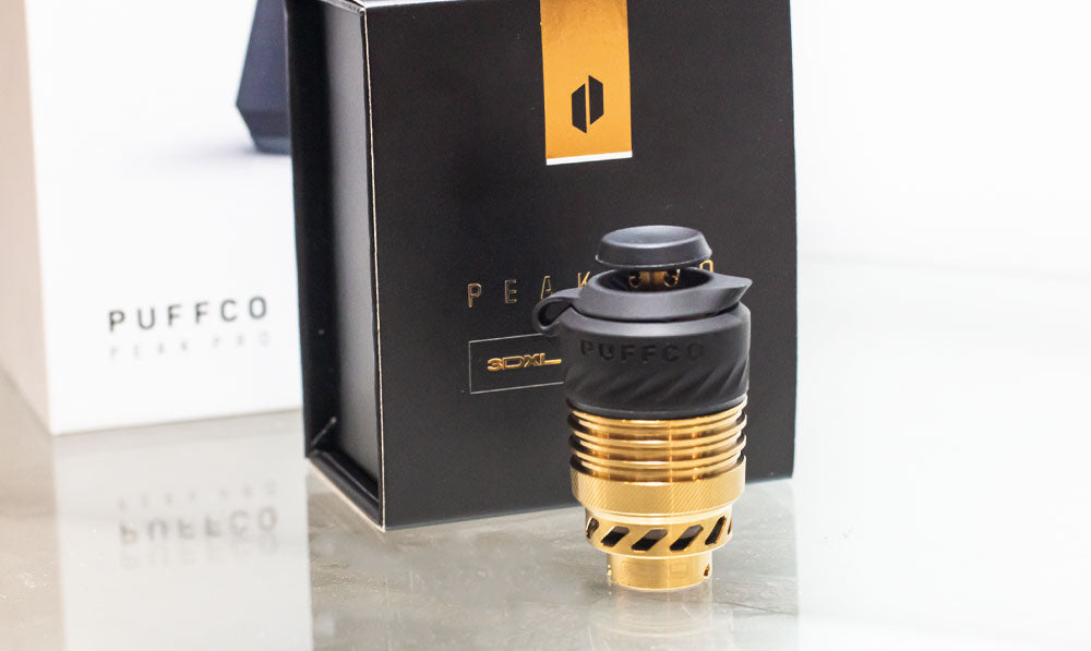 Puffco Peak Pro 3DXL Gold Atomizer resting on reflective glass surface with packaging nearby