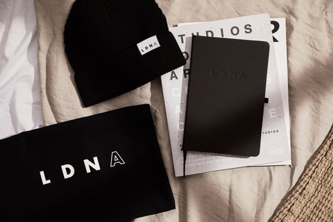 LDNA black tote bag, LDNA black note book and LDNA black fisherman cuffed beanie on a bed with magazinesen and magazine.