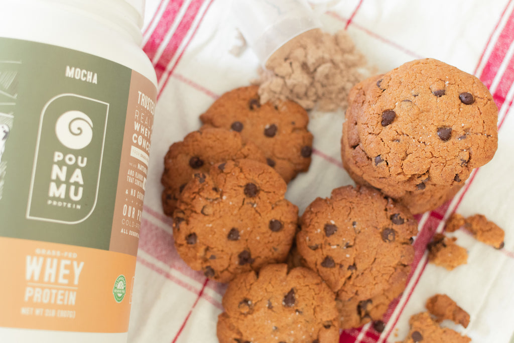 A container and scoop of Pounamu Protein Mocha Whey Protein Powder with a stack of freshly baked cookies on a red and white tea towel.