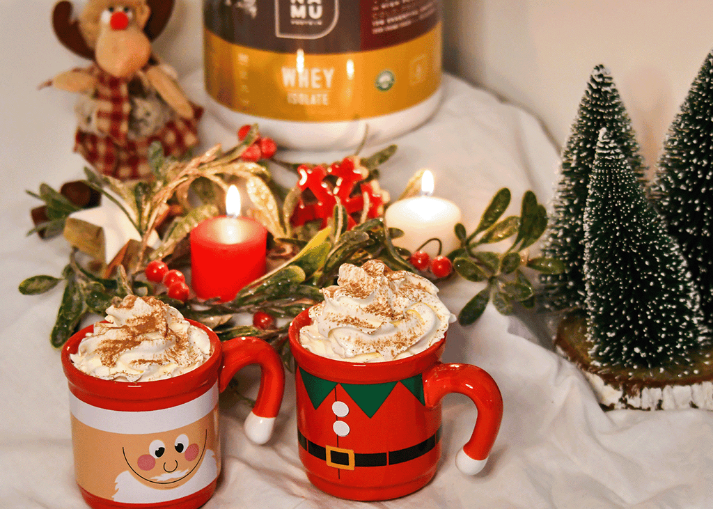 Santa and elf mug full of protein hot chocolate with whip cream on top. Candle, mini trees and wreath Christmas decorations in the background.