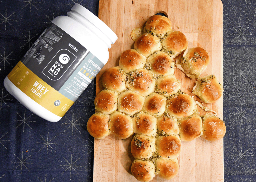 Pounamu Protein Christmas tree pull apart bread, made of 25 round buns on a wooden board on top of a blue star Christmas tablecloth with a container of Pounamu Whey Protein Isolate. 