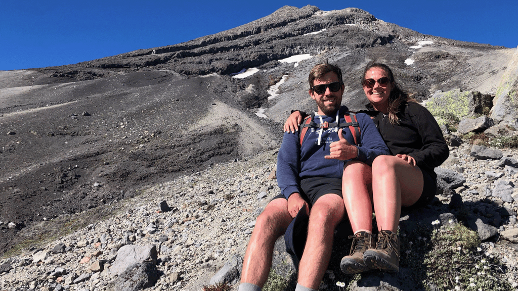 Pounamu protein staffer Alison with her finance, sitting on top of a rocky mountain with blue skis in the background.