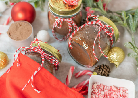 Protein powder hot chocolate mix in containers as a stocking stuffer with holiday decorations, crushed candy canes, and a scoop of protein powder