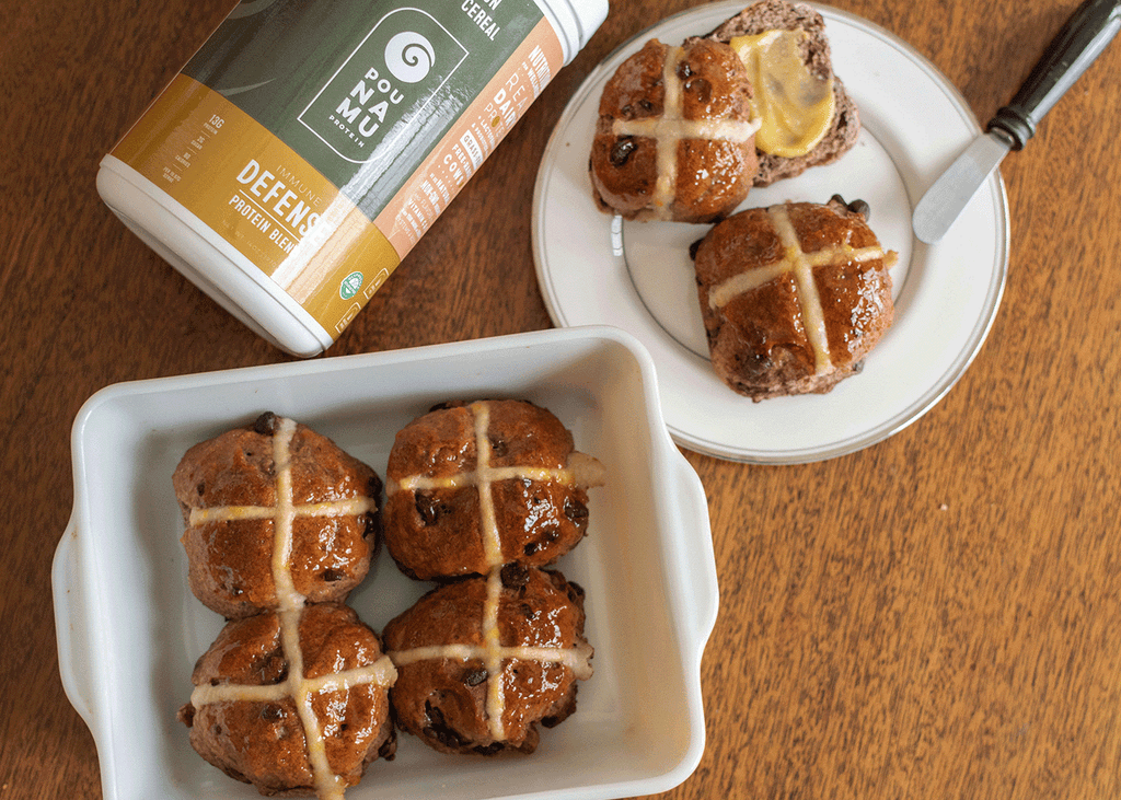 Freshly baked protein hot cross buns with chocolate chips, one is cut in half with a dollop of butter. Made using Pounamu Protein Cinnamon, Honey and Cereal Immune Defense Blend.