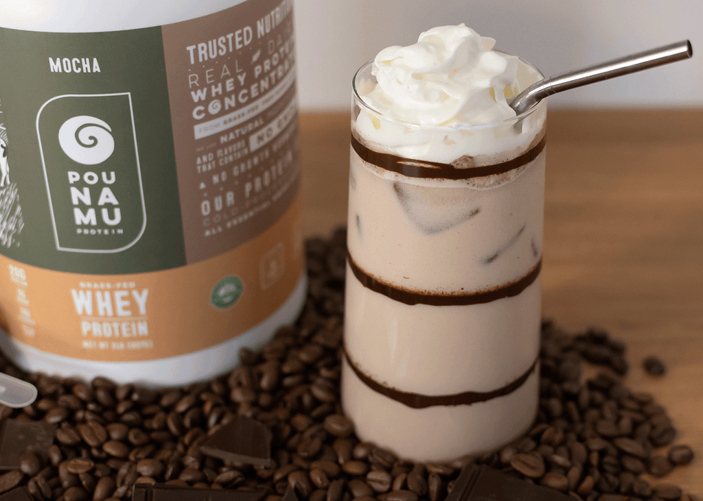 Pounamu Protein Iced Mocha in a glass with chocolate stripes and whip cream in pile of coffee beans and chocolate.