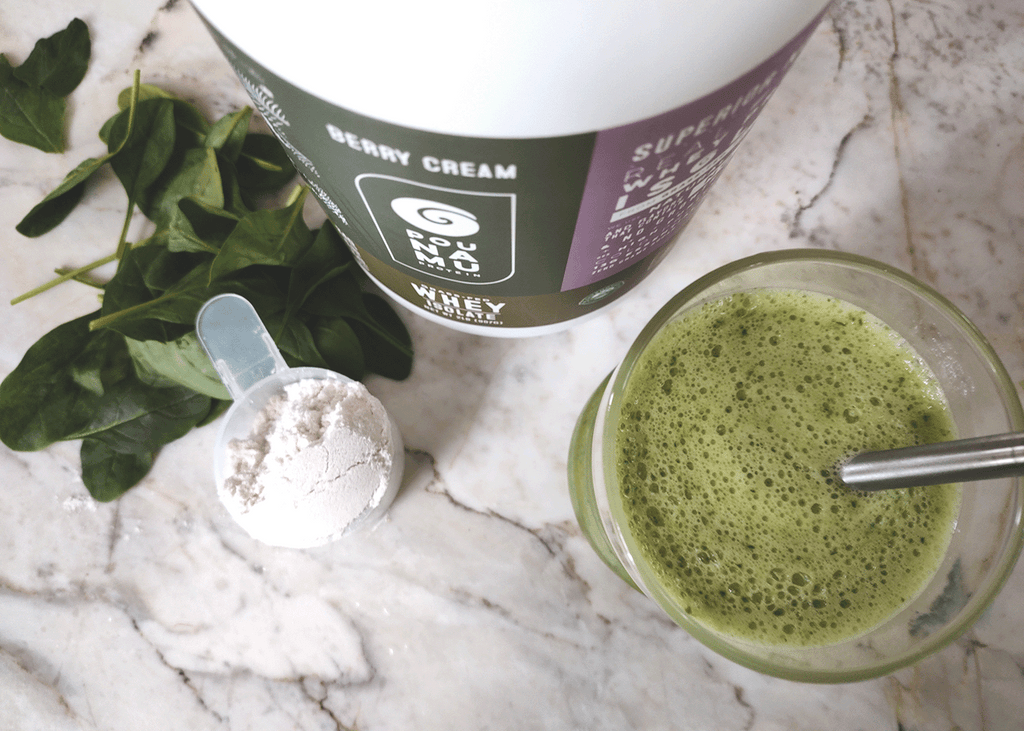 View from the top of the Pounamu spinach banana protein shake in a glass with a container of Berry Cream Whey Protein Isolate and a pile of fresh spinach on a marble background.