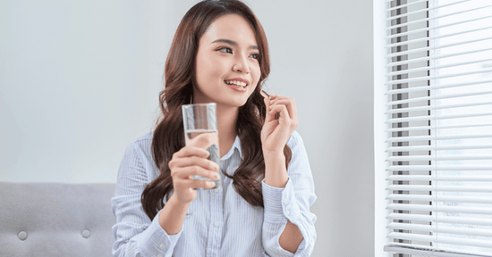 A woman holds a glass of water and a vitamin while looking out the window.