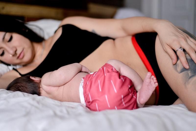 A mother in her 4th trimester breastfeeds her baby lying on her side.