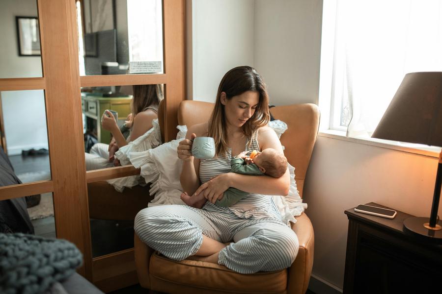 Une femme est assise dans un fauteuil avec bébé dans les bras et tasse à la main.