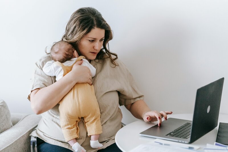 A woman in her postpartum period works at the computer with her baby in her arms.