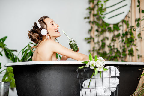 Young relaxed woman listening to the music and drinking smoothie while lying in the bathtub