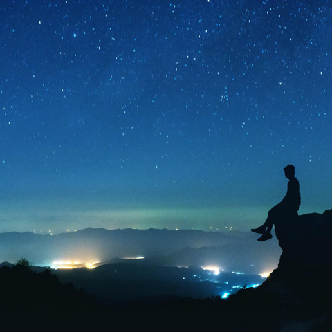 A man sitting atop a cliff at night, immersing in the cosmic energy of the universe.