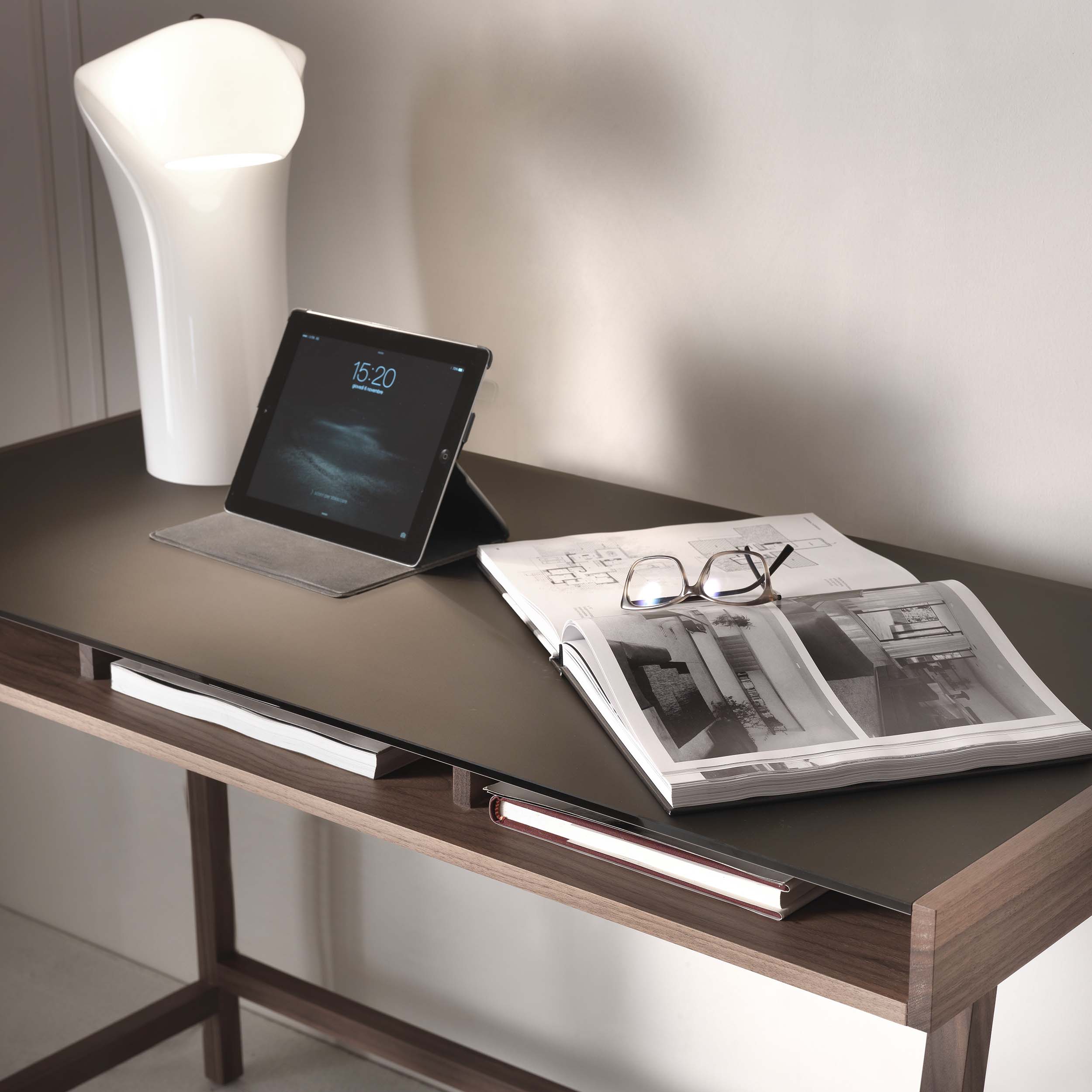 Close-up of the Abaco desk with an open book, eyeglasses, and a tablet resting on the surface, accompanied by a modern white lamp.