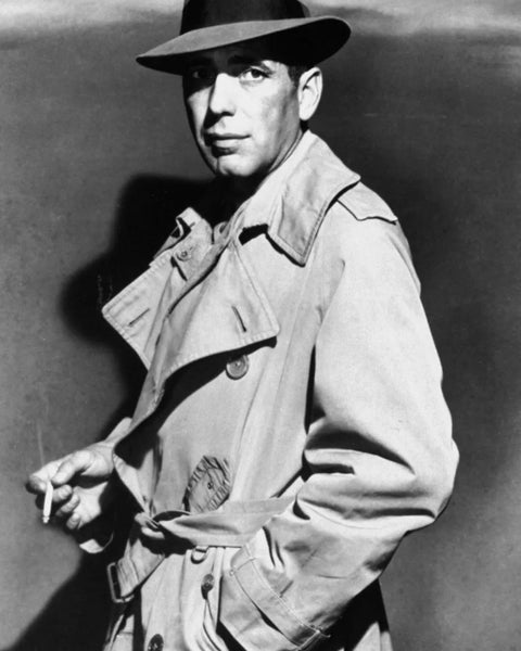 Black and white photograph of a white man standing sideways looking beyond the camera. He wears a light burberry trench coat and a trillbey hat.