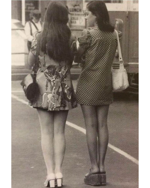 Black and white portrait photograph of two women in vintage mini skirts standing on an urban road with their backs to the camera.