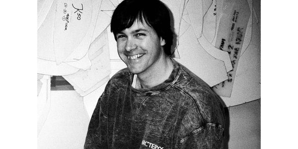 Black and white image of a white man smiling. He has dark hair that sweeps across his face and he's wearing a grainy Arctyerx sweatshirt. In the background is a wall of papers.