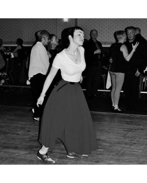 Black and white photograph of a single woman dancing in a dance hall. She has cropped dark hair and wears a light t-shirt and a-line skirt. People in the backgroun also dancing.