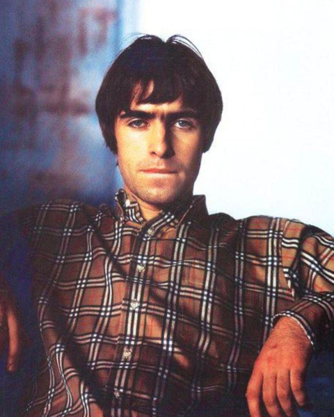 Photograph of a white man with sweeping brown hair. He's relaxing with his arm leaning on something out of sight. Wears a Burberry shirt made up of beige background and red, black and white lines