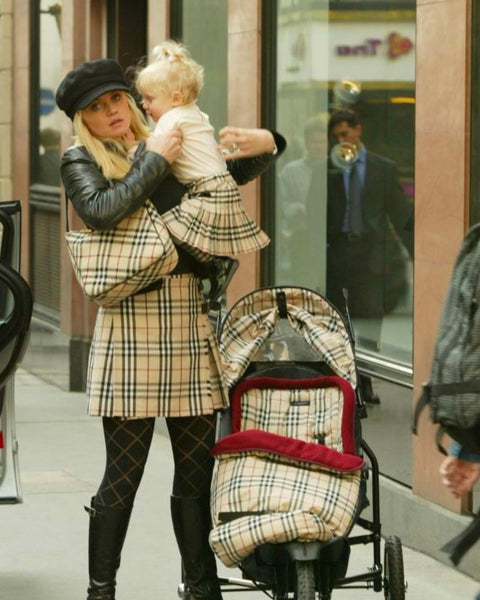 A photograph of a woman on the street. She's picking up her baby with her stroller next to her leg. She wears a Burberry skirt and bag, her child wears a burberry skirt and the stroller is made up of the burberry nova check print.