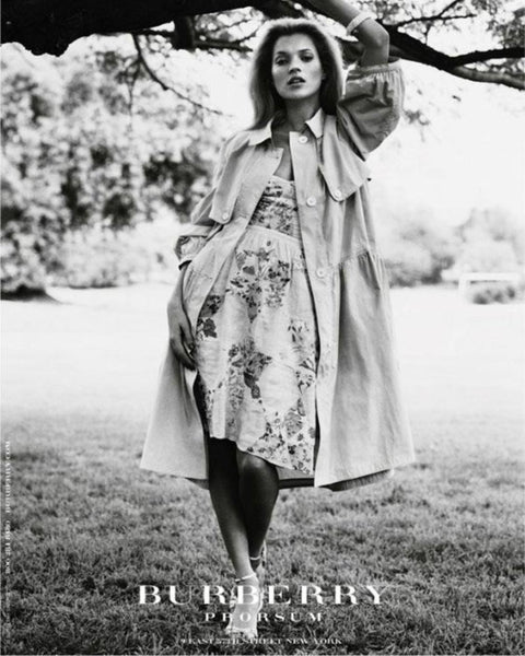 A black & white magazine advert featuring a white woman wearing a trench coat and floral, knee high summer dress. She stands in a grassy field under a tree and leans a branch with her left arm.