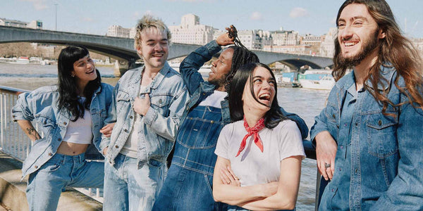 Landscape image of a mixed gender group of 5 people all wearing denim and smiling at each other. Backdrop of a London skyline.