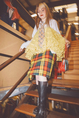 A white woman walking down the stairs wearing a white shirt and tie with a yellow tutu on top, a red green and yellow kilt and big black boots.