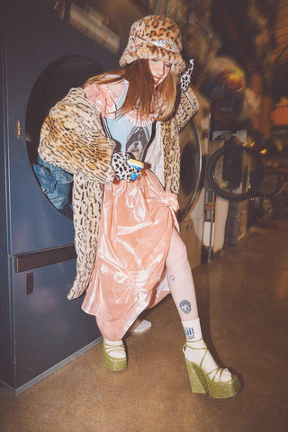 A white woman stepping out of a large washing machine wearing a pink taffeta dress, furry leopard print coat and hat, and large platform green boots.