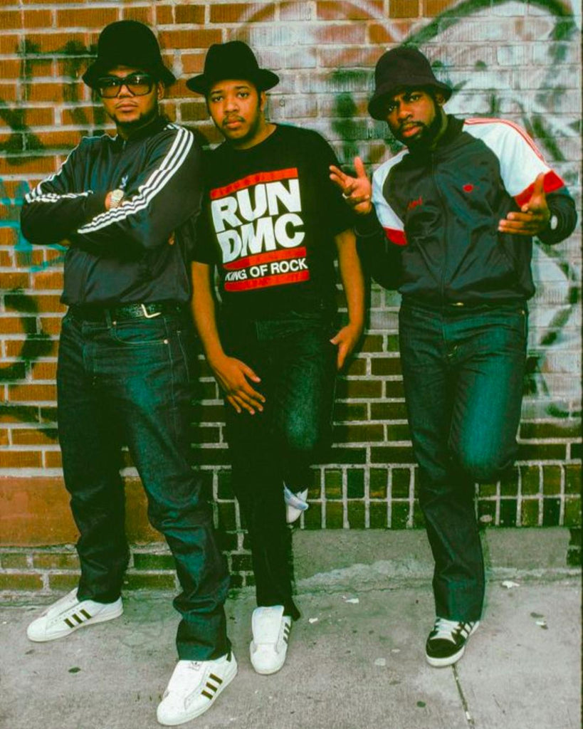 Photograph of 3 black men leaning up against a graffit'd wall. All wearing black fedora hats, two with adidas jackets and all wearing adidas trainers.