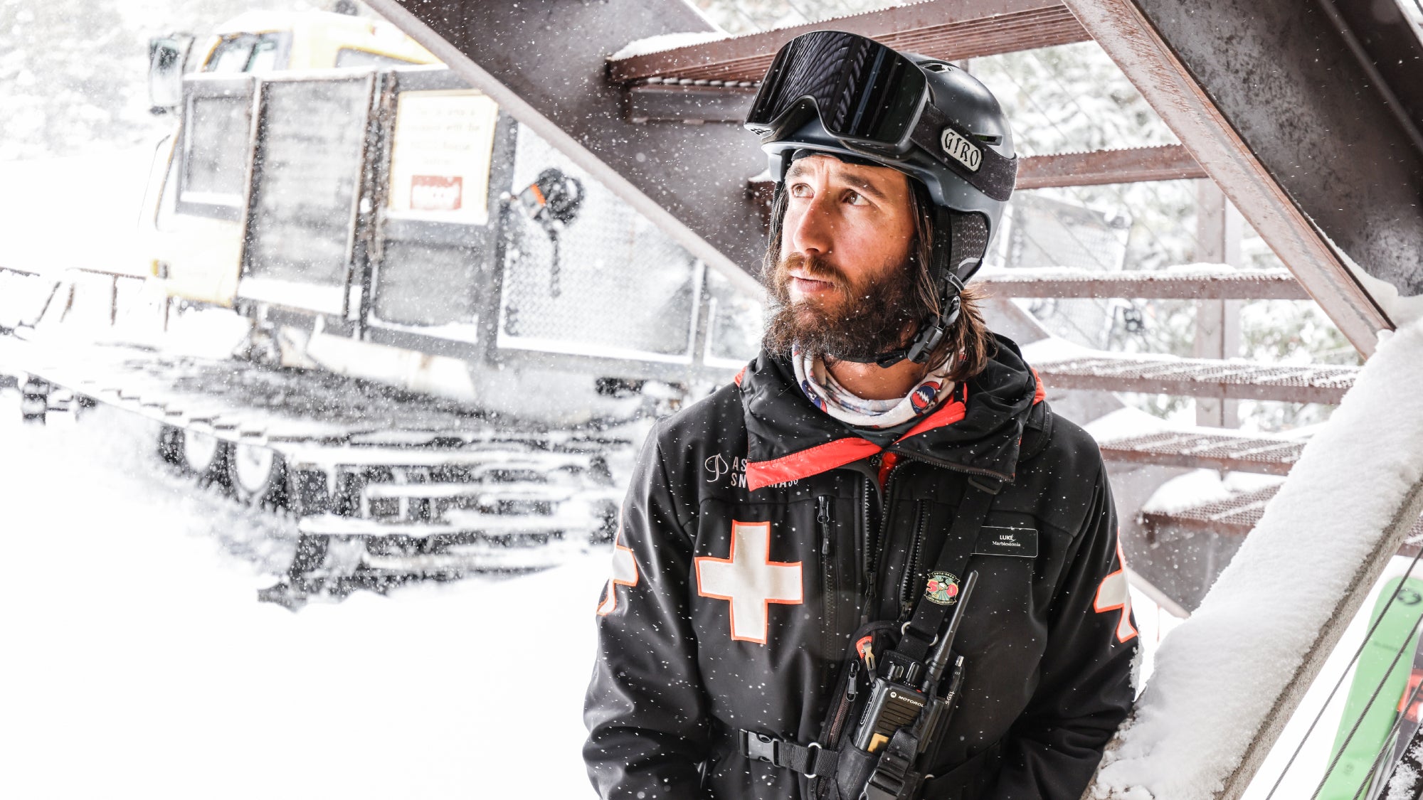 Luke Demuth standing under a stairway in heavy snow with a snowcat vehicle behind him.