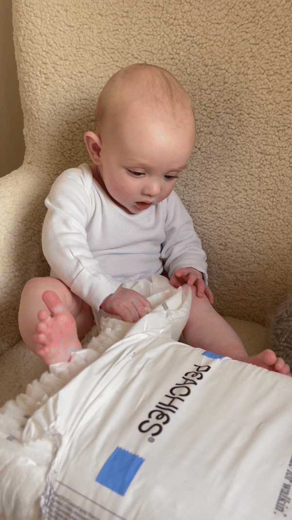 A baby in a white onesie looks at a package of diapers.