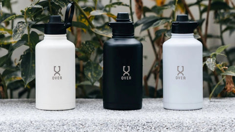 Three OVER water bottles in neutral colors lined up against a backdrop of plants, emphasizing stylish hydration solutions for active individuals.