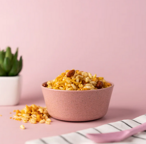 A bowl of crispy rice snack mix on a pink background with a white and pink striped napkin and a small potted plant to the side.