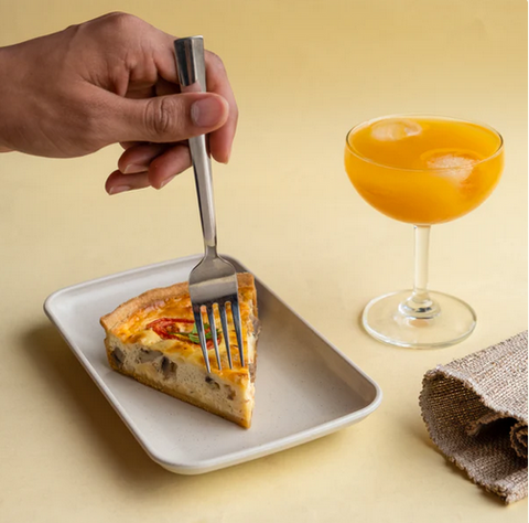 A hand piercing a slice of quiche on an eco-friendly rectangle plate, with a glass of orange juice and a burlap napkin on a yellow background.