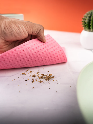 Hand using a pink sponge wipe to clean up crumbs on a marble counter, with a cactus pot and dish nearby.