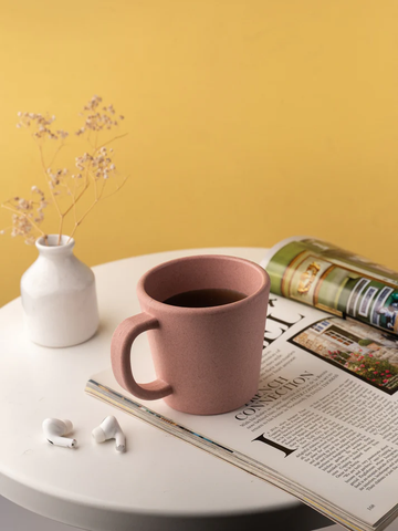 A pink kitchenware mug on a magazine with earbuds, beside a white vase with dried flowers, against a yellow backdrop.