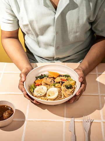 A person holding a wheat straw ramen bowl filled with noodles and mixed vegetables, with a side bowl of soup and eco-friendly utensils on a tiled table.