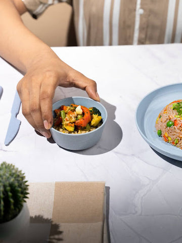 A hand reaching for a rice husk bowl filled with colorful vegetables on a marble table, with a second plate and a succulent plant nearby.