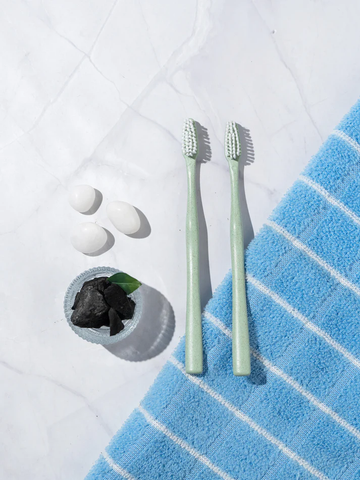 Two disposable toothbrushes on a marble surface next to a blue towel, with charcoal and stones for a clean aesthetic.