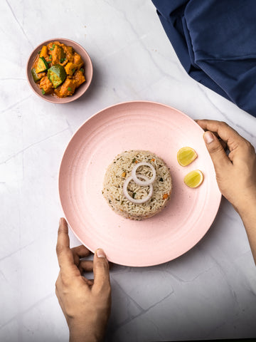 Hands holding an eco-friendly pink plate with a rice dish garnished with lemon slices, and a small bowl with a vegetable curry on a marble surface with a dark blue napkin to the side.