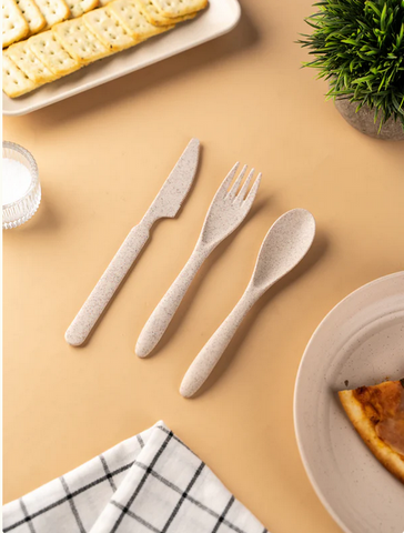 Eco-friendly cutlery set on a beige table, with crackers on a plate, a piece of pie on another plate, a checkered napkin, and a potted plant in the background.