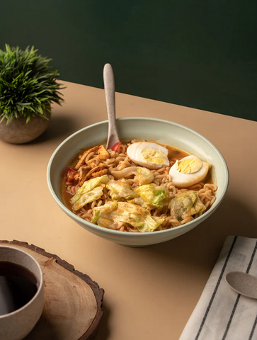 A bowl of ramen with sliced boiled eggs and vegetables in an eco-friendly bowl, with a spoon, on a two-toned table next to a cup of coffee and a cloth napkin.