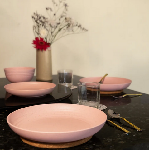 A dining table set with pink eco-friendly plates and bowls, with glasses, cutlery, and a vase with flowers and dried plants as the centrepiece.