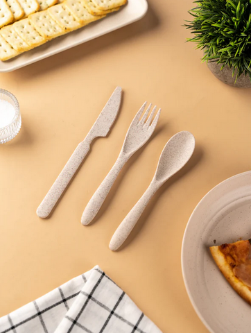 A set of eco-friendly cutlery on a beige table, with a plate of crackers, a piece of pie, a glass of water, a checkered napkin, and a potted plant in the background.
