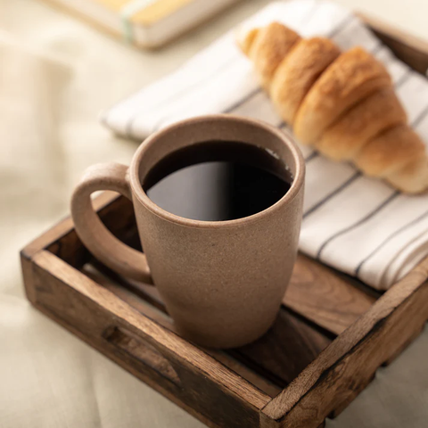 A rice husk coffee mug filled with coffee on a wooden tray beside a croissant, evoking a warm, relaxing tea or coffee time.