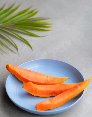 Slices of ripe papaya on an eco-friendly blue plate with a palm leaf in the background, on a grey surface.