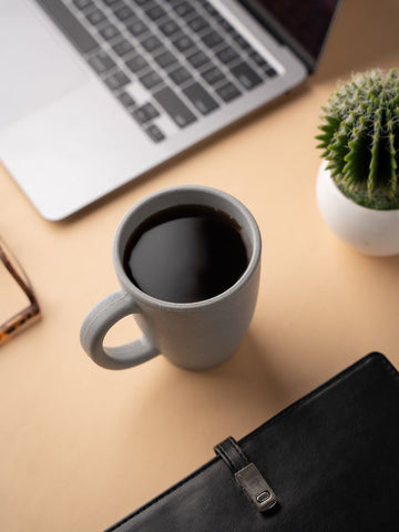 A gray wheat straw coffee mug filled with black coffee on a beige desk, with a laptop's keyboard in the upper left, a potted cactus plant in the upper right, and the corner of a black leather portfolio in the foreground.