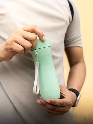 Close-up of a hand holding a mint green, eco-friendly reusable cup with a wrist strap, against a soft yellow background.