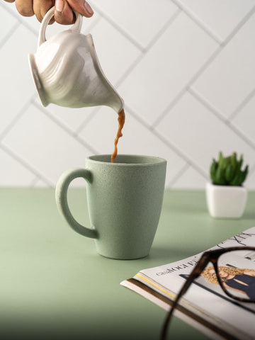 A hand pouring coffee from a white jug into a mint green eco-friendly cup on a green table, with a notebook and a small potted plant in the background.